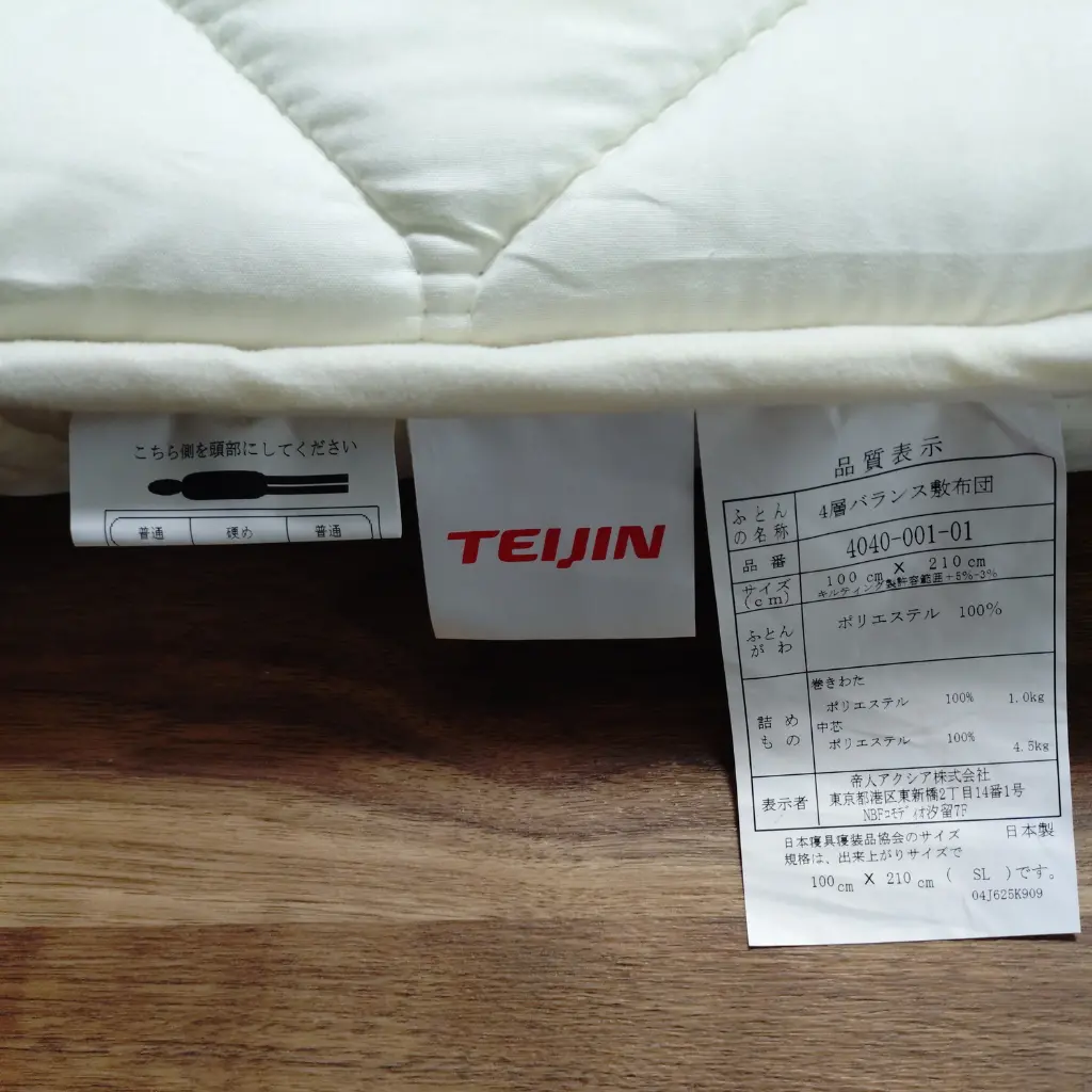 寝姿勢サポート敷布団の品質タグ写真。TEIJINロゴと素材表示、サイズ表記が写っている。